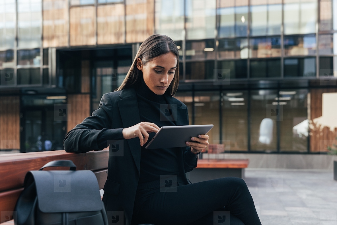 Confident businesswoman in black stylish formal attire using digital tablet Female entrepreneur sitting outside in courtyard working remotely