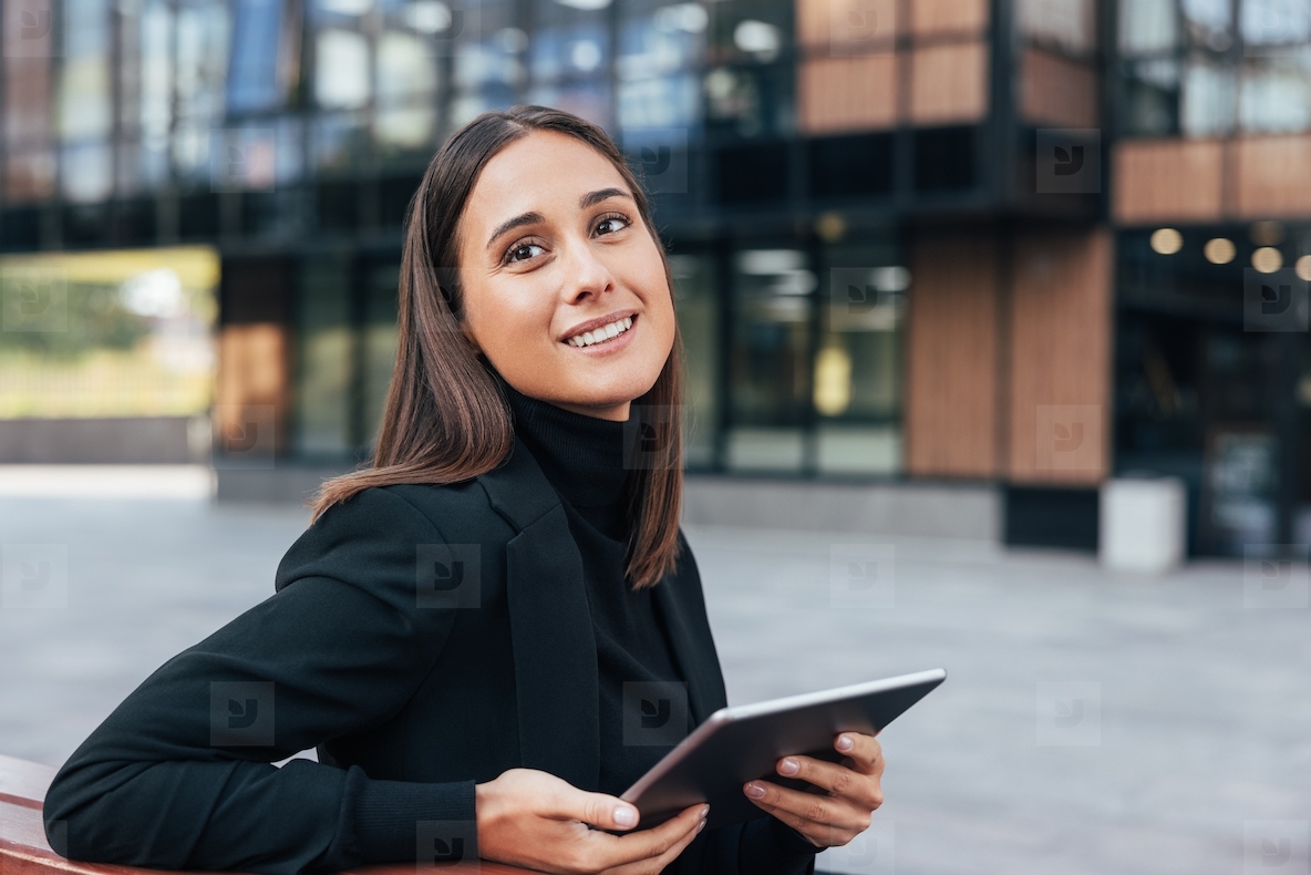 Beautiful smiling businesswoman holding a digital tablet and looking away  Young woman in black formal attire sitting outside