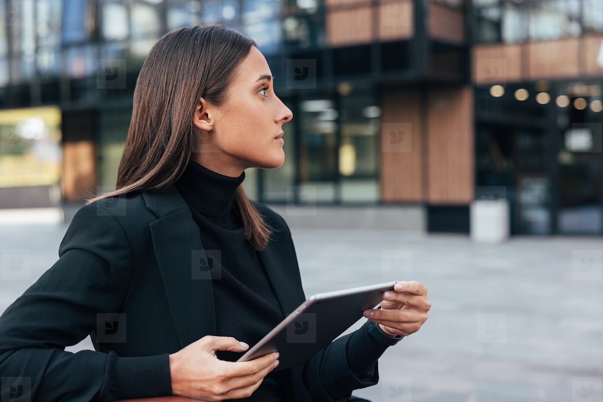Side view portrait of young businesswoman with black hair sitting outside looking away  Female with digital tablet sitting outside against office building