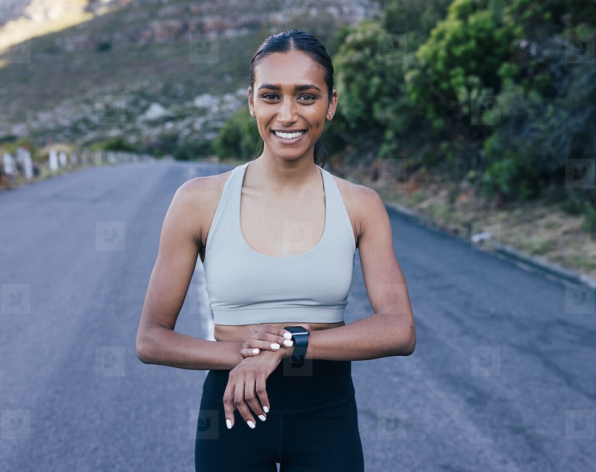 Smiling woman in fitness attire looking at camera and using smartwatches while standing on abandoned road