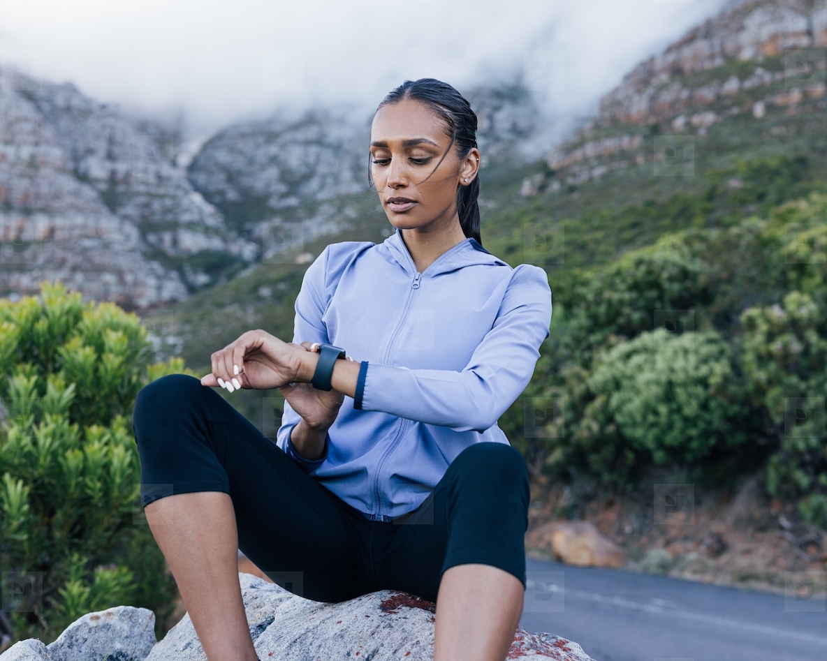Young confident sportswoman in fitness attire checking pulse while sitting on rock against mountains