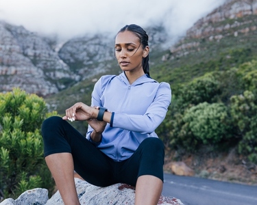 Young confident sportswoman in fitness attire checking pulse while sitting on rock against mountains