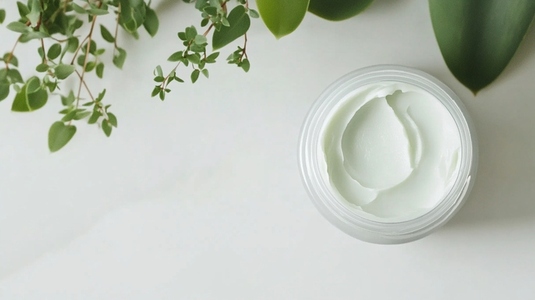 Open jar of face cream with fresh green leaves beside it
