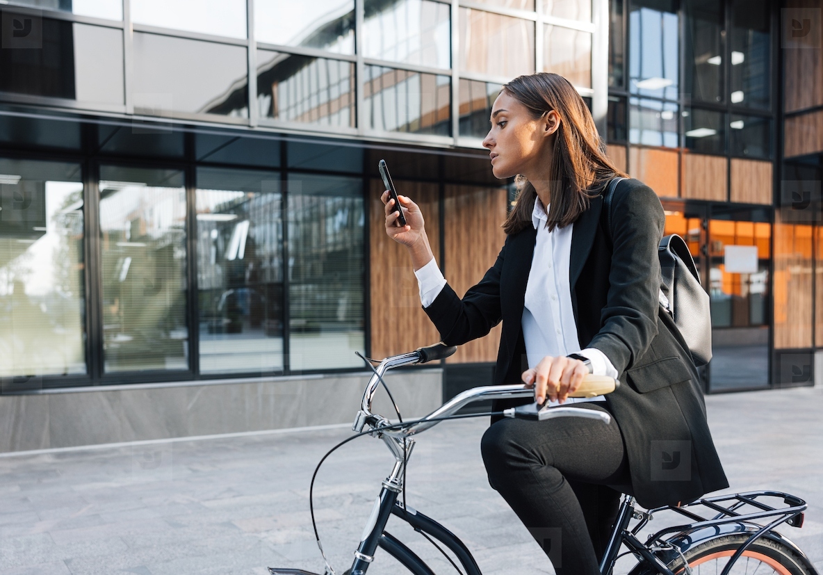 Businesswoman sitting on bicycle and checking her mobile phone  Young female in formal clothes using mobile phone while sitting on bicycle