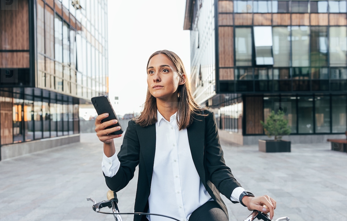 Businesswoman standing outdoors with bicycle and holding mobile phone  Portrait of a young female in formal clothes standing on the courtyard of an office building