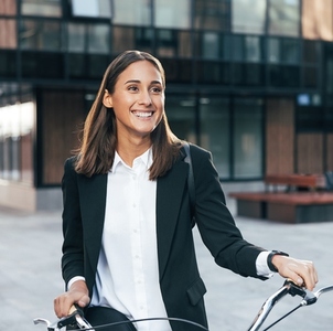 Happy female entrepreneur standing outdoors with bicycle at business building  Portrait of a smiling businesswoman wearing formal wear and a backpack holding bicycle