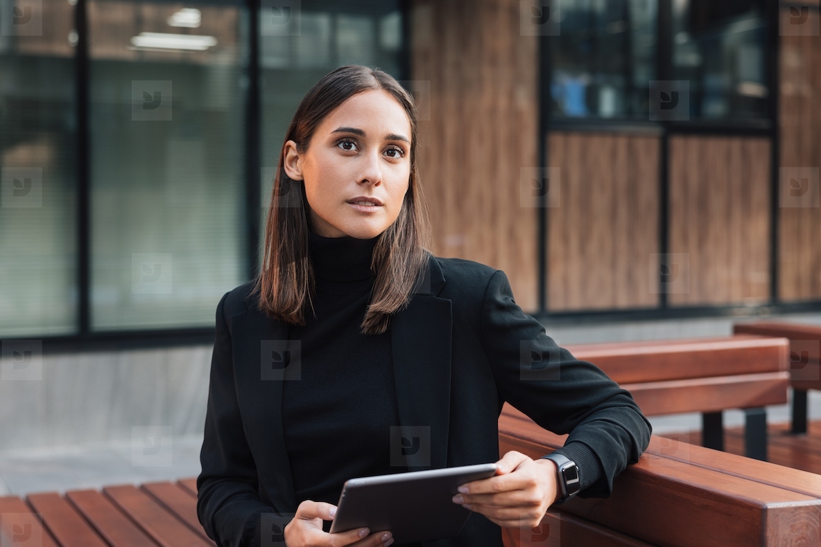 Portrait of young businesswoman in black stylish attire sitting outdoors with a digital tablet