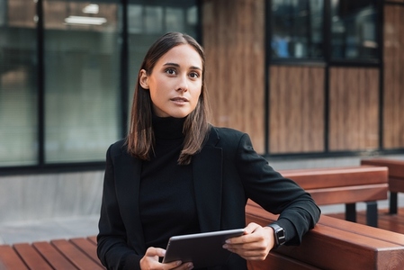 Portrait of young businesswoman in black stylish attire sitting outdoors with a digital tablet
