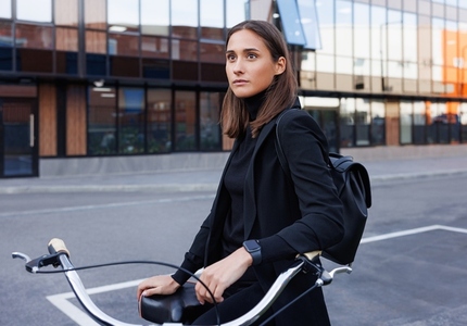 Portrait of a confident female in black formal wear wearing backpack  Stylish female entrepreneur standing outdoors with bicycle