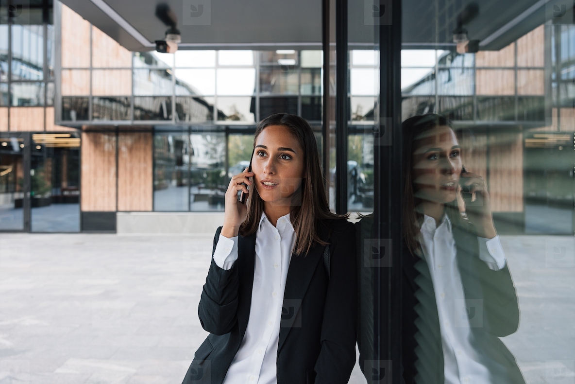 Young businesswoman leaning against a wall of a glass office building talking on mobile phone