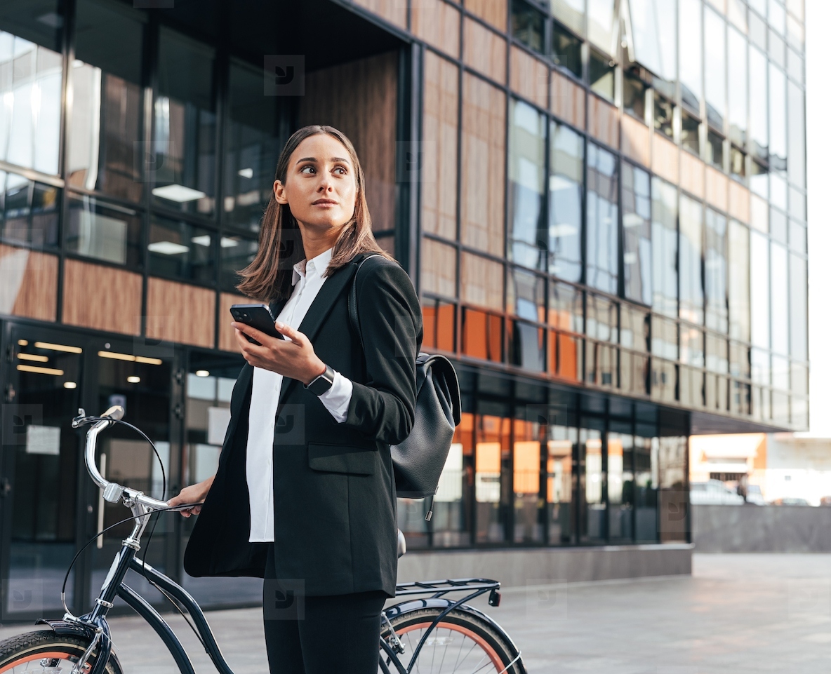 Confident businesswoman standing with bicycle and holding smartphone in the city