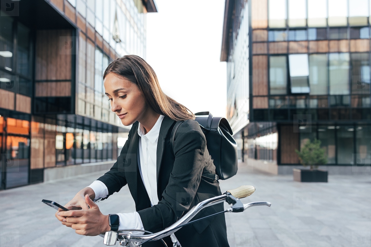 Young woman in fromal wear leaning bicycle and checking her mobile phone