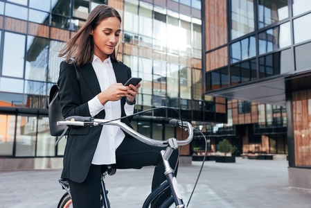 Young businesswoman sitting on a bicycle and checking her smartphone  Positive female entrepreneur with bicycle in courtyard of business building