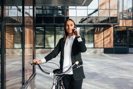 Confident female in formal wear with bicycle talking on mobile phone  A young female entrepreneur is talking on a mobile phone while standing with a bicycle in the courtyard of a business building