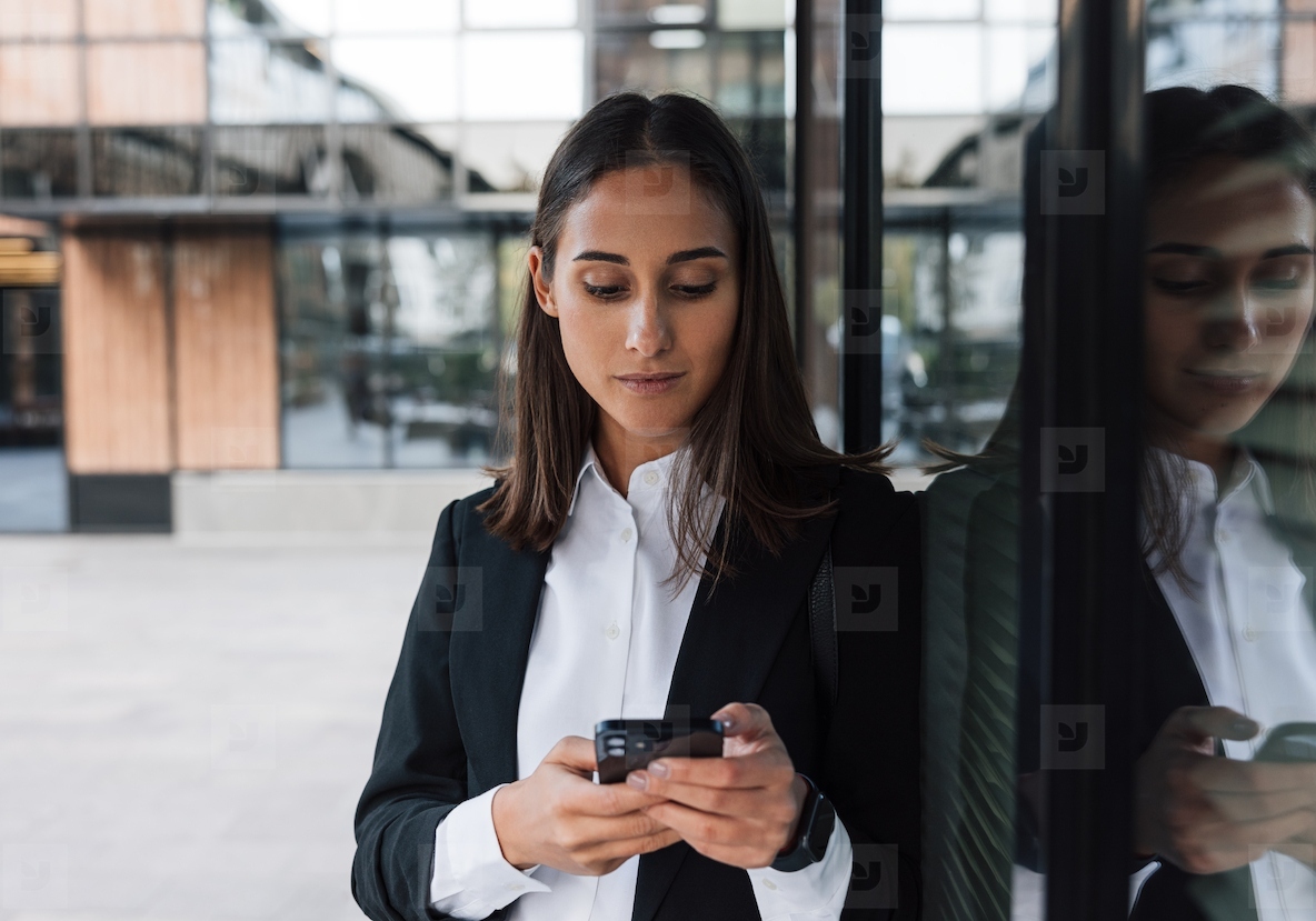 Portrait of a young female entrepreneur using a smartphone while leaning against the glass of a business building