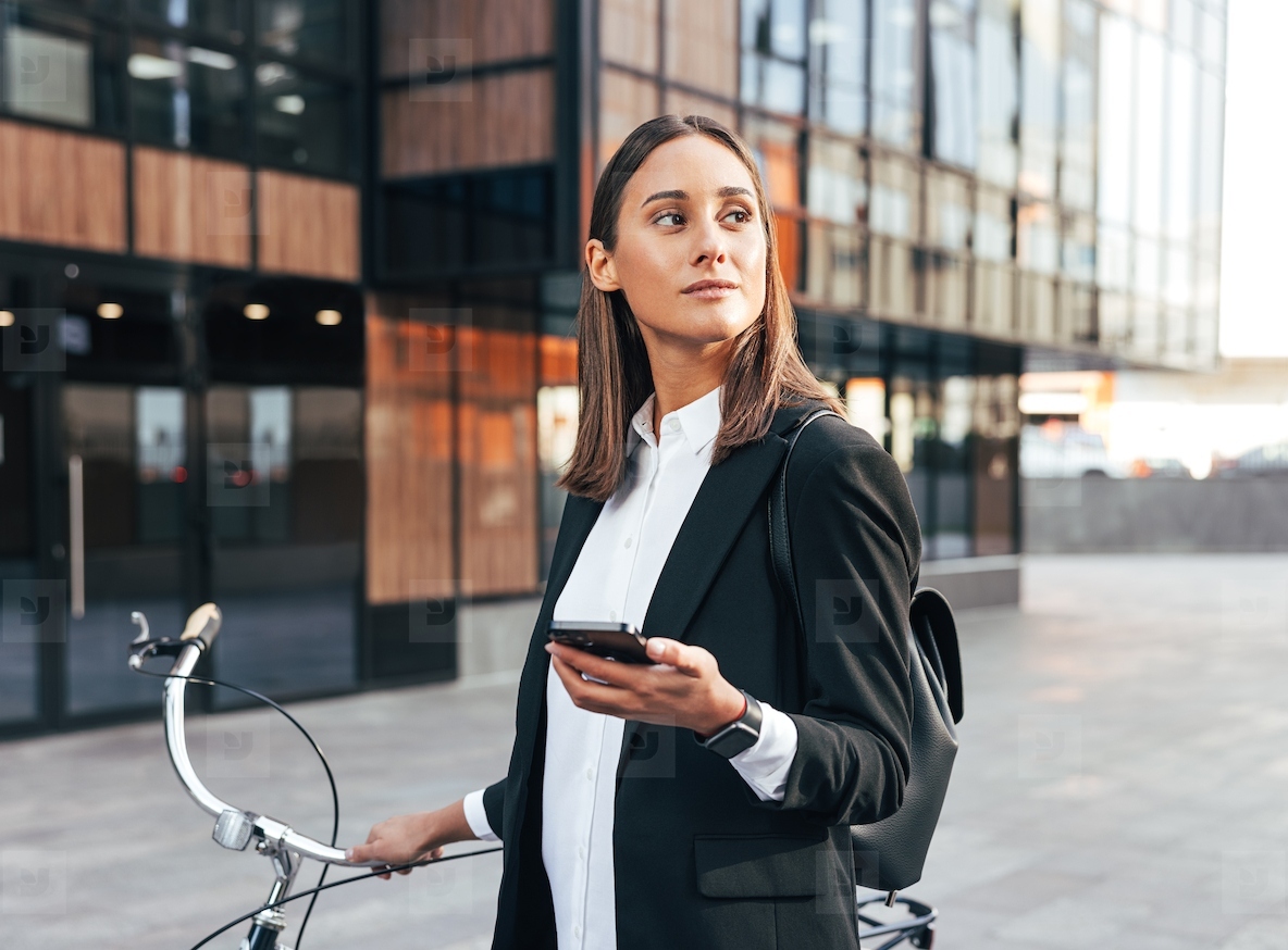 Young confident businesswoman with mobile phone and bicycle standing outdoors wearing formal wear