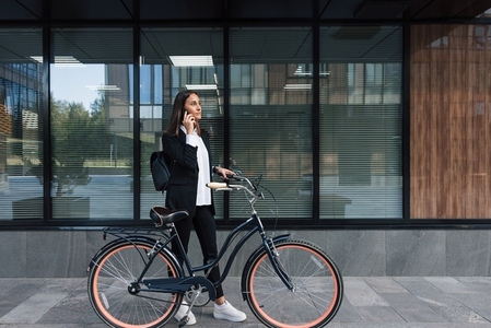Businesswoman standing with a bicycle at the office building taking on mobile phone Female in formal clothes is making a phone call during a bicycle ride Businesswoman standing with a bicycle at the office building taking on mobile phone Female in formal clothes is making a phone call during a bicycle ride