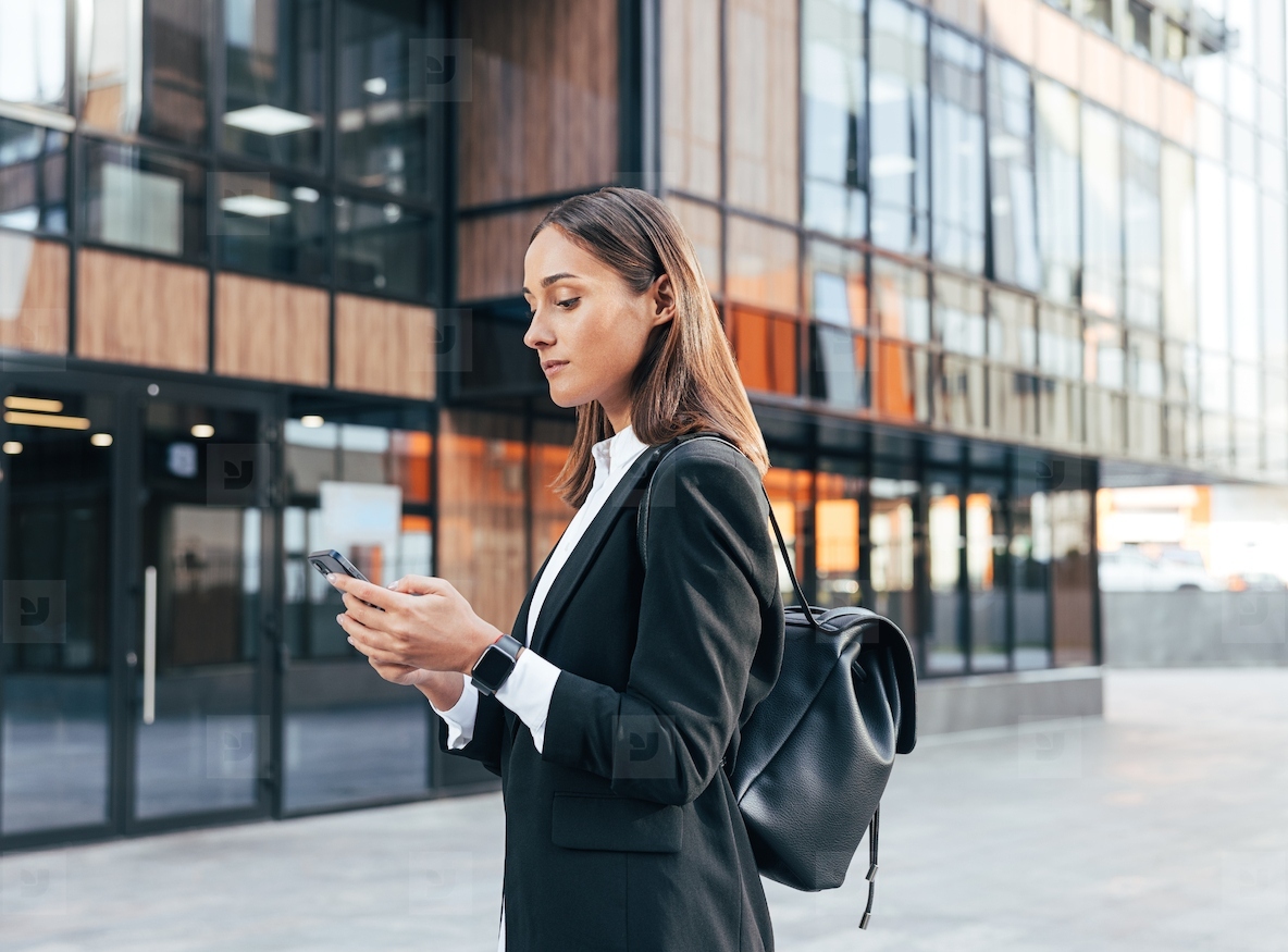 Female in black formal clothes wearing backpack typing on mobile phone while standing against an office building