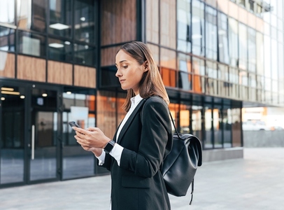 Female in black formal clothes wearing backpack typing on mobile phone while standing against an office building