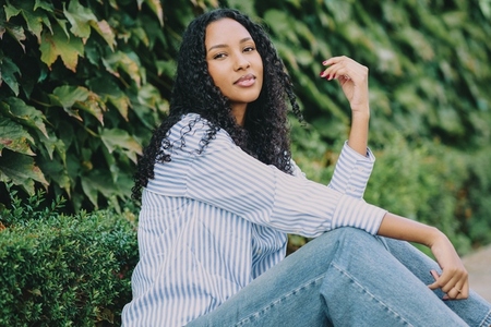 A Stylish Young Woman Sitting Comfortably Against a Backdrop of Lush Greenery in Casual Attire A Stylish Young Woman Sitting Comfortably Against a Backdrop of Lush Greenery in Casual Attire