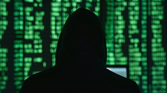 hooded figure stands before glowing green code symbolizing cyber threats hooded figure stands before glowing green code symbolizing cyber threats