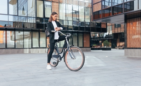 Young woman in business wear checking her watch while standing with a bicycle in the courtyard of an office building  Female in black office wear during her ride to work