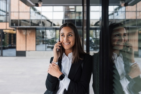 Smiling female in black formal wear leaning business building and talking on mobile phone  Female office worker talking on mobile phone during a break