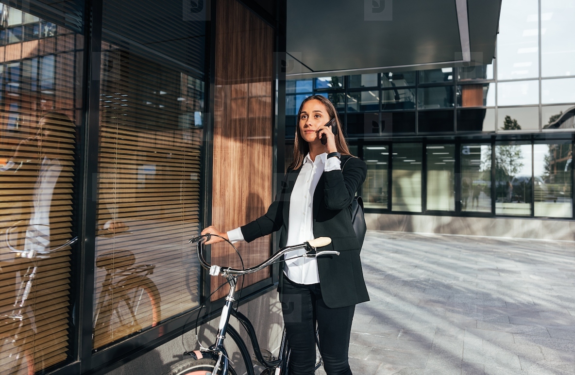 Young female entrepreneur talking on mobile phone while walking outdoors with bicycle Woman in formal wear is talking on mobile phone while standing outdoors at a business building