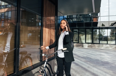 Young female entrepreneur talking on mobile phone while walking outdoors with bicycle  Woman in formal wear is talking on mobile phone while standing outdoors at a business building