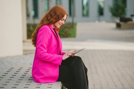 A Professional Woman Dressed in a Stylish Pink Blazer Engaging with a Tablet Outdoors in the City