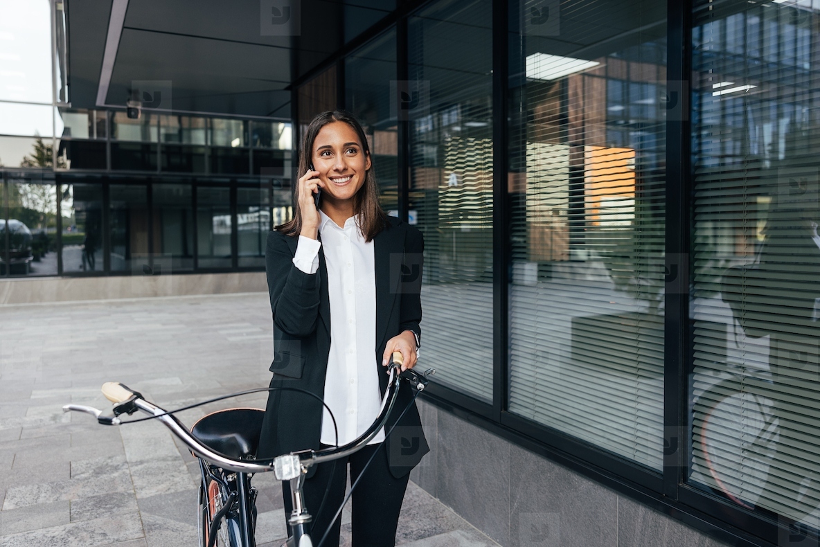 Smiling confident female in formal wear walking near an office building with a bicycle and talking on mobile phone