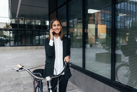 Smiling confident female in formal wear walking near an office building with a bicycle and talking on mobile phone