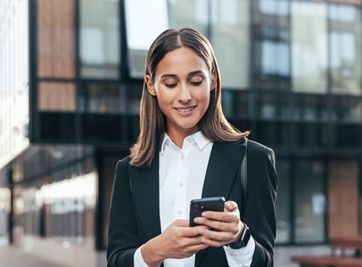 Smiling confident woman in formal wear on the way to work  checking her phone  standing in the courtyard of an office building