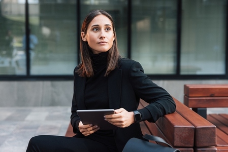 Serious and stylish woman sitting outdoors with a digital tablet and looking away