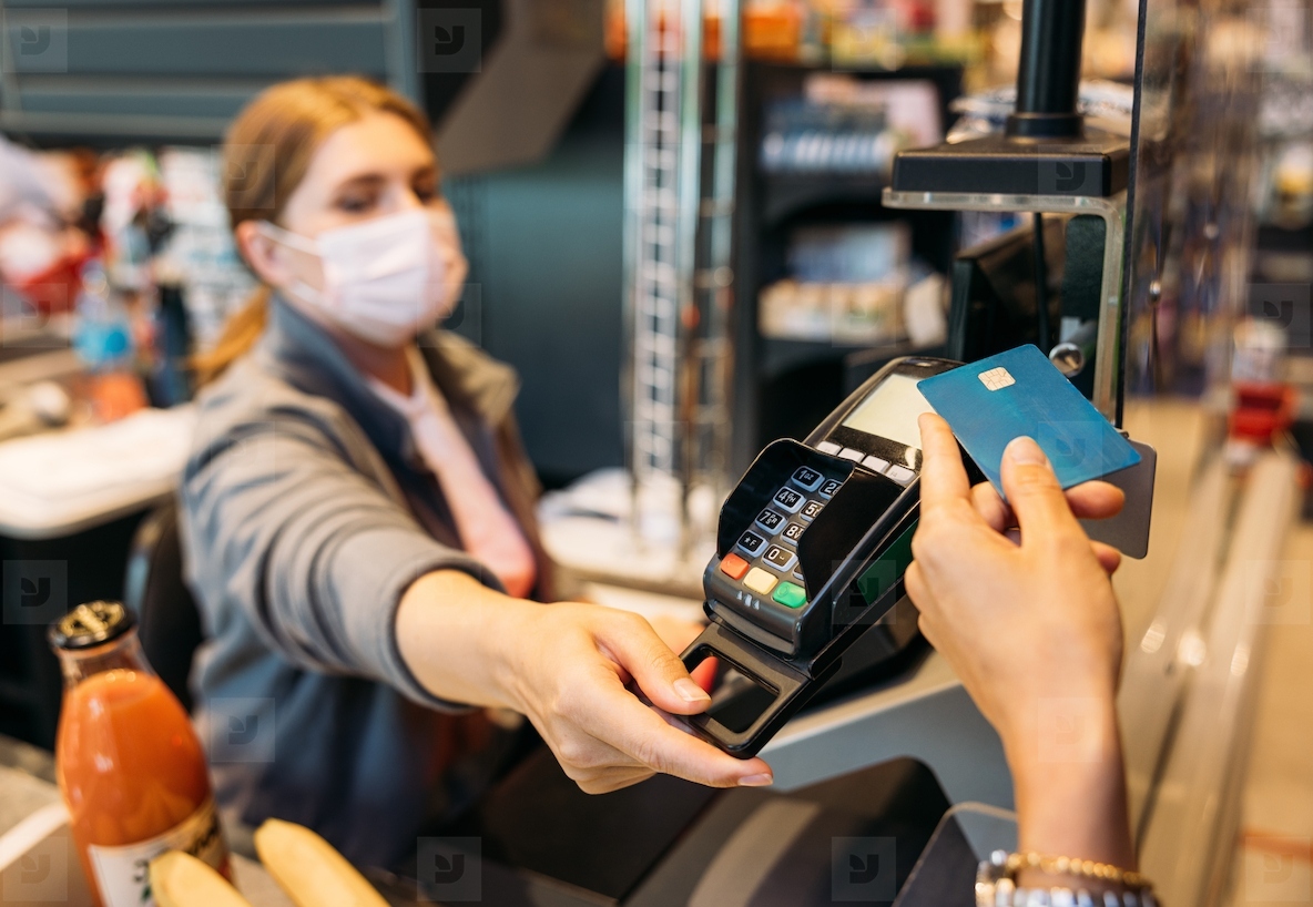 Unrecognizable womans hand holding a credit card with NFC chip while the cashier in a mask turns the payment terminal towards the customer