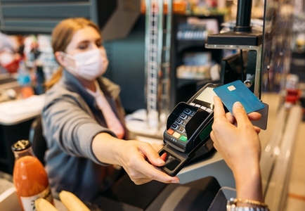 Unrecognizable womans hand holding a credit card with NFC chip while the cashier in a mask turns the payment terminal towards the customer