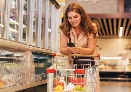 Smiling female customer leaning on the cart and checking her smartphone