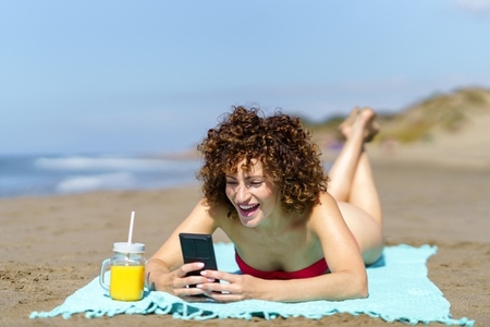 Laughing woman using smartphone while lying on beach