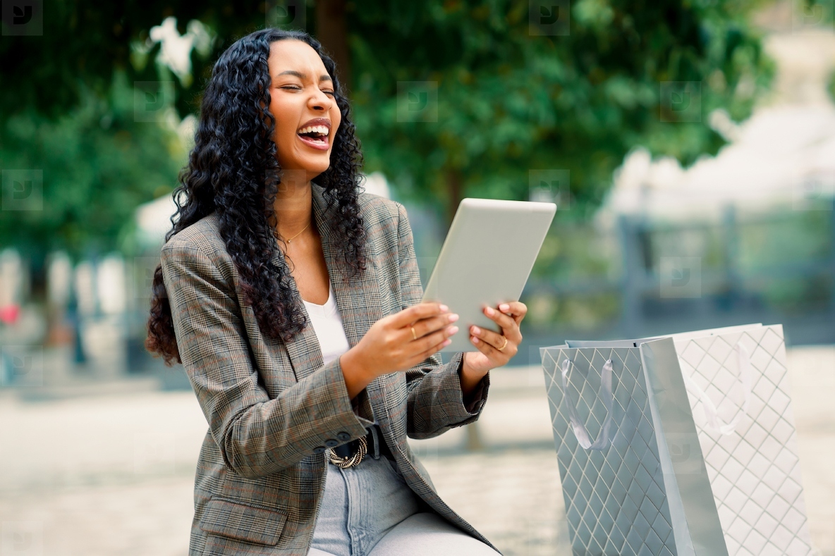 A Smiling Woman Delightfully Enjoys Using a Tablet While Relaxing in an Urban Park Setting