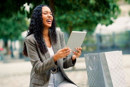A Smiling Woman Delightfully Enjoys Using a Tablet While Relaxing in an Urban Park Setting