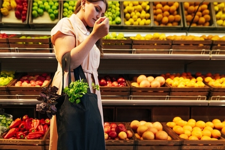 Young woman holding a bag with vegetables in her bent hand  standing at shelves in a grocery store
