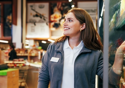 Young smiling female grocery store worker standing at the fridge and looking away