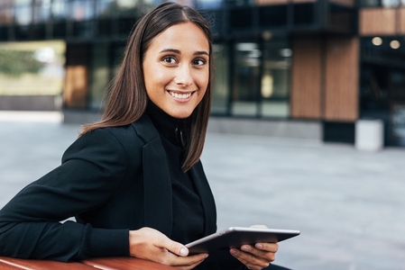 Smiling brunette woman in black formal clothes sitting outdoors with digital tablet