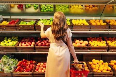 Rear view of a woman in casual clothes standing at shelves with fruits and vegetables holding shopping basket