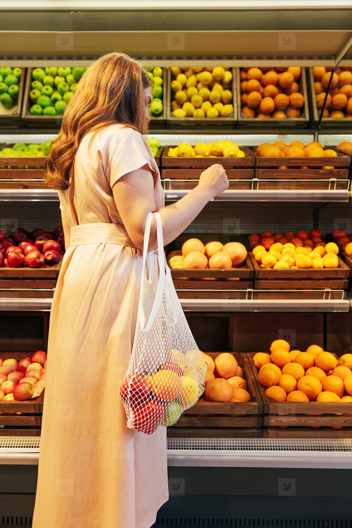 Back view of young woman choosing fruits while standing at shelves in grocery store