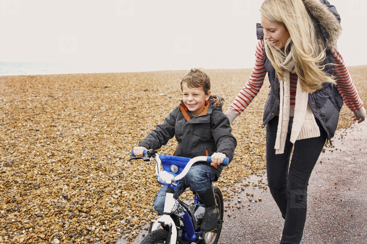 Happy mother helping son learn how to ride bicycle on path along rocky beach