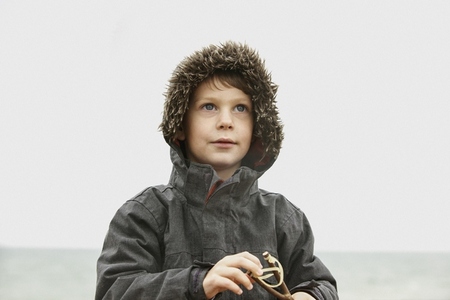 Curious boy in fur lined jacket holding slingshot on winter beach