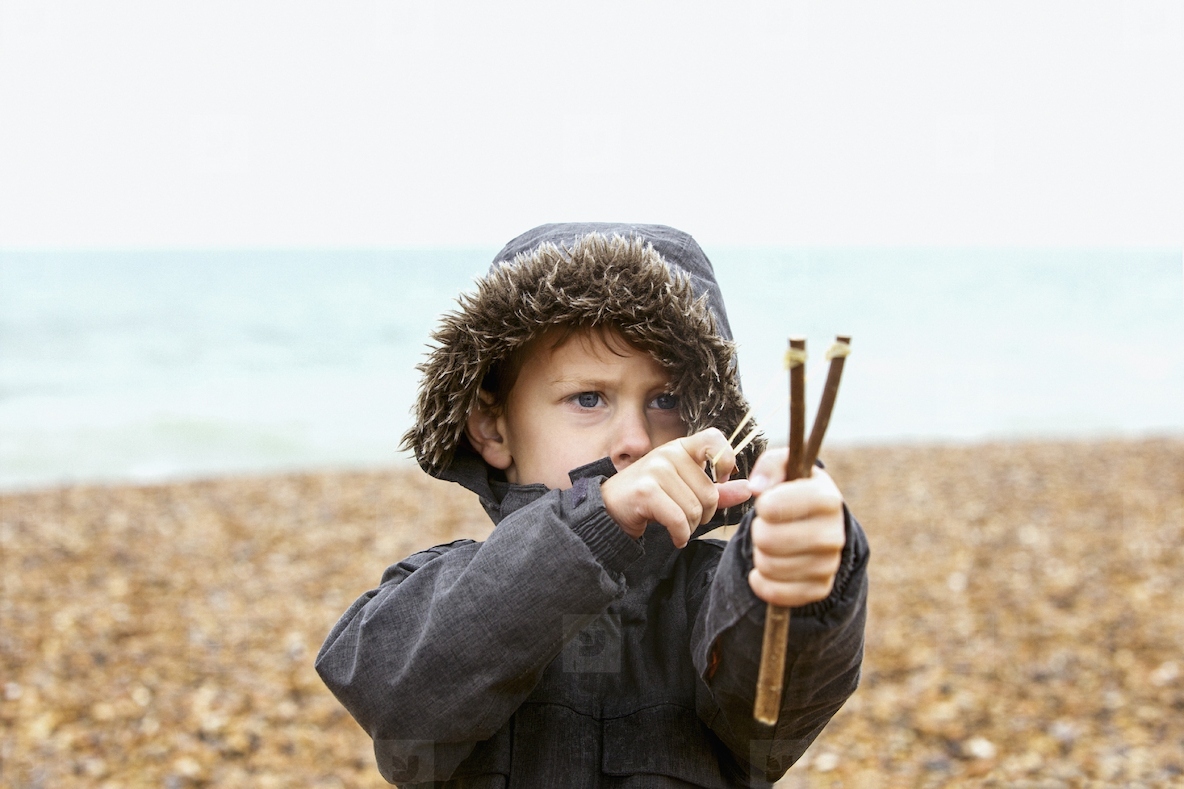 Cute boy in fur lined jacket playing with slingshot on rocky beach