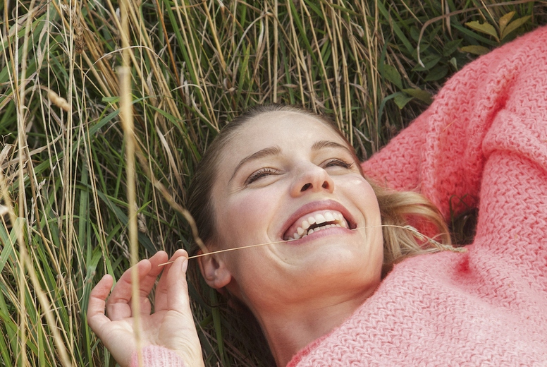 Carefree woman in pink sweater smiling and laying in tall summer grass