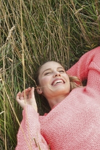 Carefree woman in pink sweater smiling and laying in tall summer grass